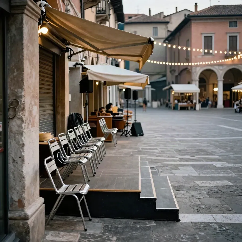 Piazza italiana al crepuscolo con sedie pieghevoli, palco e luci sospese sotto gli archi.